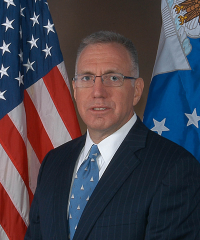 A headshot of Robert Chandler Swallow in a navy suit and light blue tie infront of an United States flag and Unitied States Air Force flag